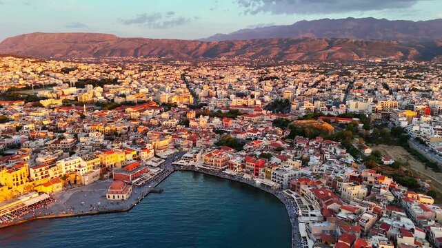Chania Old Venetian Harbor and historic waterfront extend beneath the White Mountains, with Pachnes peak rising beyond the city in Crete, Greece, drone forward flyover.