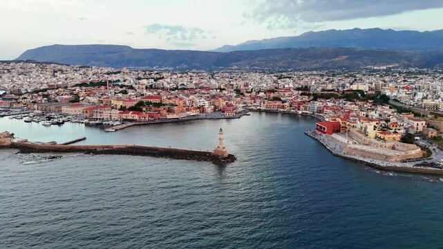Chania Old Venetian Harbour features the Egyptian Lighthouse, stone breakwater and historic waterfront beneath the White Mountains in Crete, drone flyover.