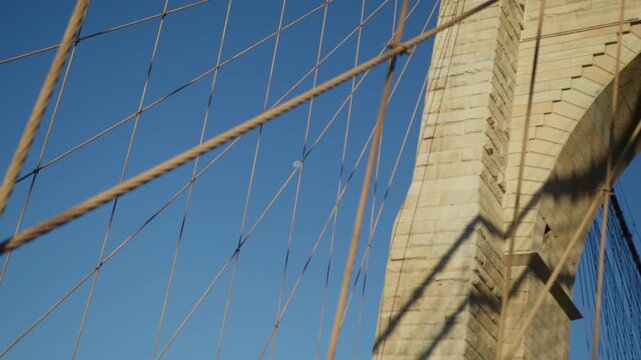 Web of steel suspension cables of Brooklyn Bridge crosses a granite bridge tower with the pale daytime moon visible through the ironwork, hard sunlight raking the stonework.