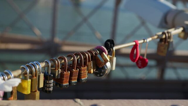 Dozens of padlocks in different colours and states of rust hang clipped to a slim bridge rail, water and mesh soft behind, warm side light on the metal.