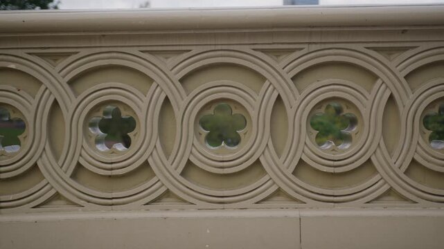 Camera tracks along a carved stone balustrade, interlocking circles and quatrefoil openings revealing glimpses of green foliage behind in soft overcast light.