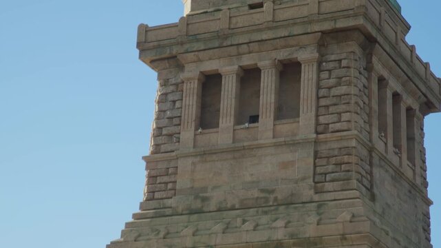 Camera tilts up the rusticated granite pedestal of the Statue of Liberty, past a band of carved stone discs to the shaded colonnade recesses at the top, clear blue sky behind.