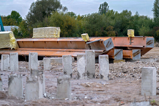 Concrete foundation piles protruding from dirt ground with large orange metal girders and structural beams stored at an industrial construction site.