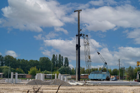 Heavy construction pile driving rig positioned on open land surrounded by precast concrete piles under a cloudy blue sky.