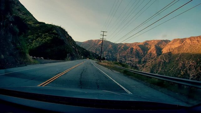 A dashcam view of Malibu Canyon Road winding through mountain landscape with utility poles lining side of road under clear sky during golden hour