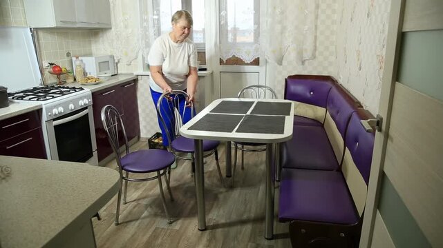 Elderly woman arranges chairs around small dining table in kitchen interior, preparing for meal or gathering, home environment, daily routine, domestic activity, household task.