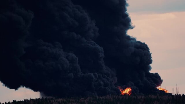 Massive industrial fire sending a thick column of black smoke into the sky. A catastrophic explosion at a petroleum product storage facility causes dangerous air pollution and environmental damage