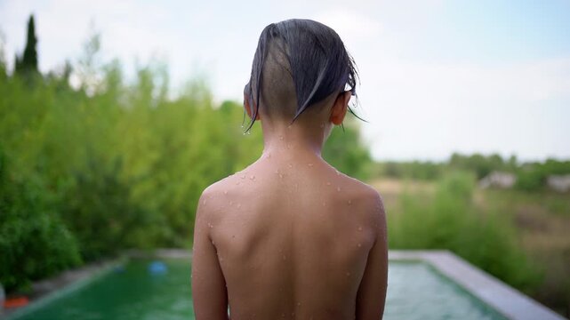 Filmed from behind, a wet boy stands looking at the swimming pool before jumping in, leaving a splash