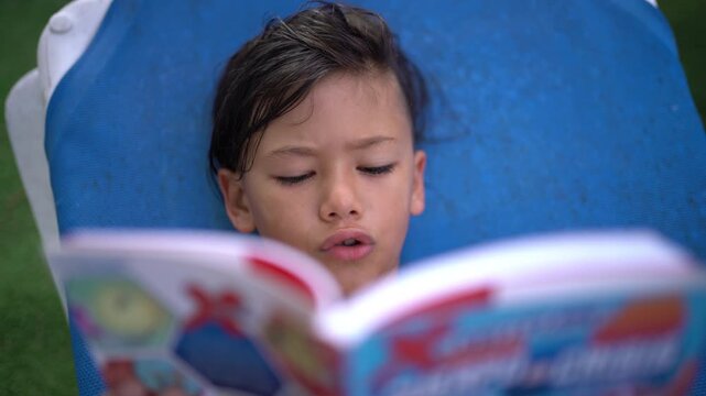 A boy with wet hair reads a comic book while lying on a blue pool lounge chair
