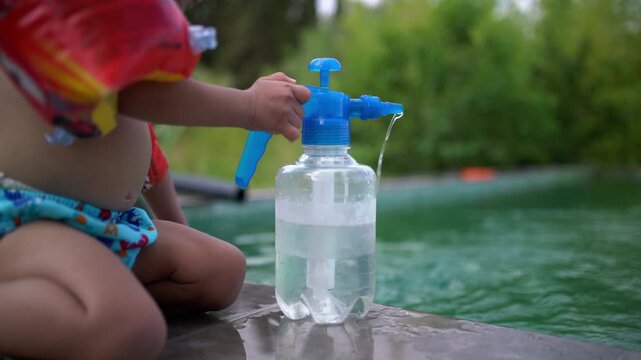 A toddler wearing inflatable arm floaties plays with a water pump bottle at the edge of the swimming pool