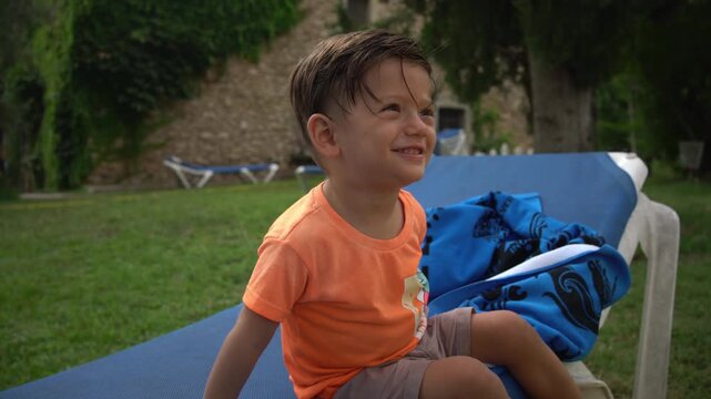 A toddler sits on a lounge chair making funny mouth sounds and laughing, with the farmhouse in the background
