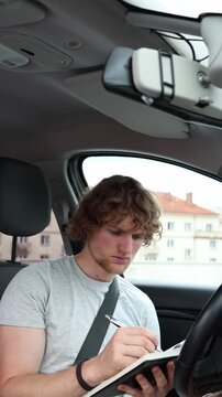 Vertical screen driver sits inside a parked car writes in a notebook and solves traffic rule questions while preparing carefully for his future driving theory examination
