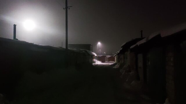 Winter night scene along snowy alleyway. Small buildings covered snow. Diffused light source streetlamp illuminates path. Cold desolate atmosphere urban winter landscape. Low visibility fog.