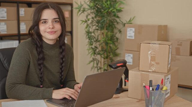 Woman typing on laptop, smiles while surrounded by packages in a building; confidence small business growth.