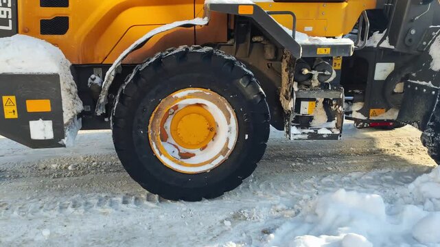 Yellow heavy equipment loader on snow-covered road with tire tracks under bright winter sunlight. Machine clears snow outdoors on sunny day with visible snow piles.