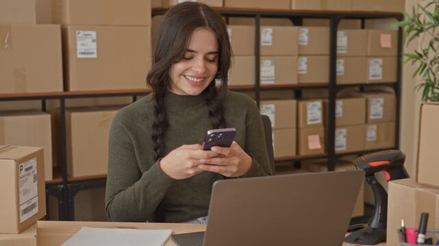 Woman holding smartphone with hands, smiling and tapping while seated at laptop among stacked shipping boxes in building; small business optimism.