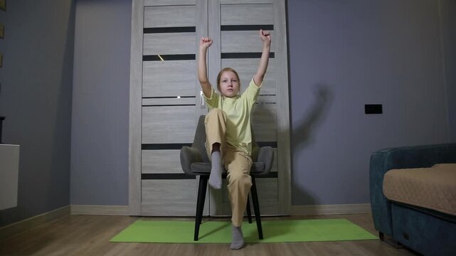 Young girl performing seated exercises indoors at home, raising knees and arms in rhythmic motion on a chair, demonstrating fitness activity for children wellness and healthy lifestyle.