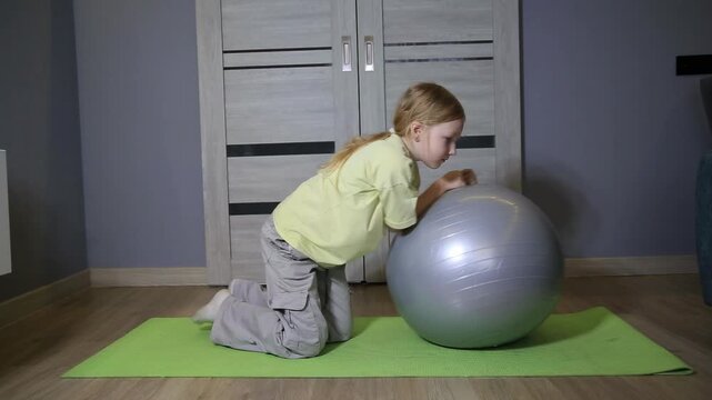 Little caucasian girl on green mat exercising at home with fitness ball during day. Child performing physical activity on wooden floor of living room. Healthy lifestyle development.