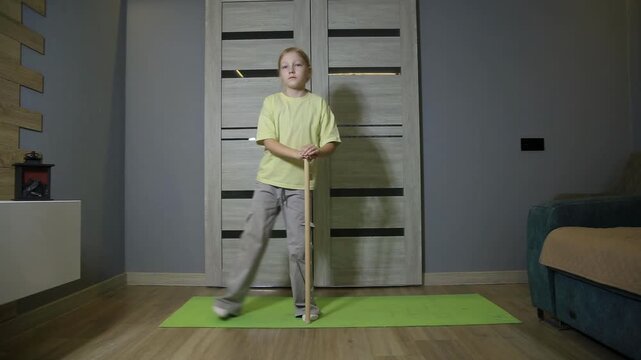 Blonde hair girl standing on green yoga mat indoors holding wooden stick preparing for exercise. Home workout of one person in living room with grey wall and modern doors.