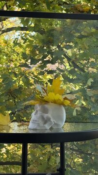 White skull planter filled with maple leaves stands on balcony table surrounded by autumn trees. Halloween balcony styling, small space seasonal decor, gothic autumn aesthetic, foraged leaf decorating