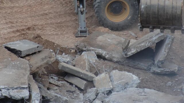 Excavator bucket moving broken concrete slabs and debris at construction site with large tire of heavy machinery on dirt ground, demolition work in progress, outdoor scene