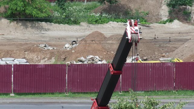 Close-up detail of heavy industrial crane boom and complex pulley system angled upwards against background of active construction site with earth piles, red barrier fence under bright sky copy space