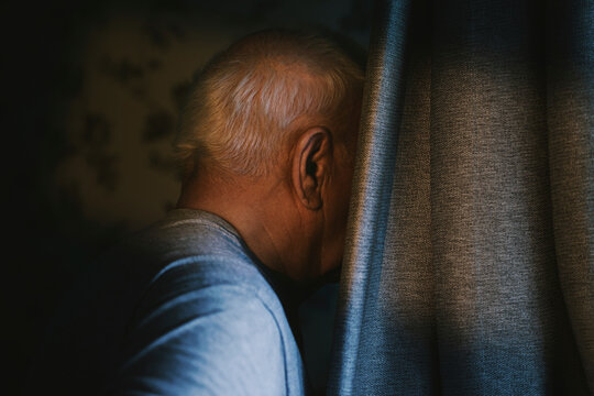 A senior man hides his face behind a dark curtain indoors