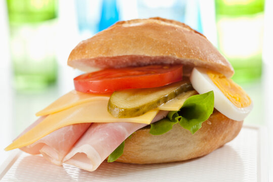 Bread roll with sliced processed cheese, ham, tomato, boiled egg and cucumber on chopping board, close up