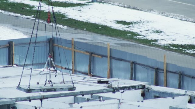 Crane lowering concrete slab onto building foundation at winter construction site, urban development project showing work progress in snowy environment