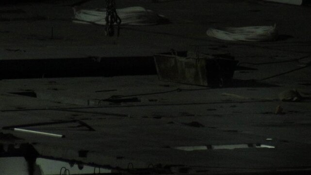 Dark industrial construction scene at night featuring hanging metal chains, rusted container, coiled cables, and debris on concrete surface