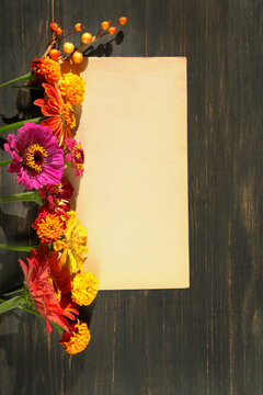 bright colorful flowers of zinnias and marigold (tagetes) and old paper sheet blank on dark wooden table close up. floral beautiful image. seasonal summer end and autumn flowers. top view. copy space