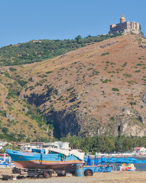 Sanctuary of the Black Madonna of Tindari perched high on mountain cliff seen from Oliveri beach with traditional blue fishing boat on shore in Marinello, Messina, Sicily, Italy.