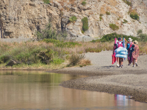 Group of tourists carrying beach gear walking along sandy shore of Laghetti di Marinello natural reserve lagoon with limestone rock wall backdrop in Oliveri, Tindari, Sicily, Italy.
