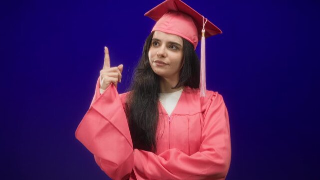 Woman in pink graduation gown points index finger upward in studio with mortarboard tassel visible and arms crossed; confidence success.