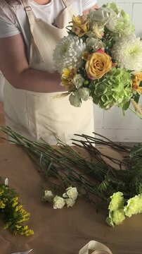 Woman florist in apron holds bouquet with roses, chrysanthemums and hydrangeas at workshop table. Small business floristry, handmade bouquet craft, flower shop workflow, artisan entrepreneurship
