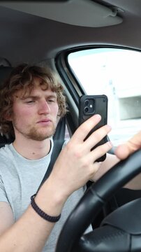Vertical screen young inexperienced driver checks a smartphone while driving through traffic showing poor judgment and creating a dangerous risk for everyone on busy road