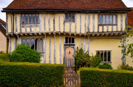Medieval half-timbered house with yellow limewashed walls, exposed oak beams, arched Gothic windows and a wooden door behind a clipped hedge in Lavenham, Suffolk, England
