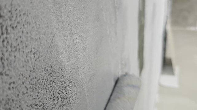 a paint roller applying white paint coat onto a rough concrete wall, showcasing detailed tactile surface texture for home renovation, interior design, DIY wall painting, and construction concepts.