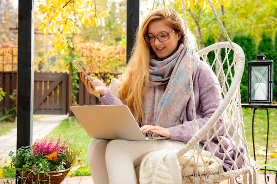 A mid adult woman works remotely on a laptop while drinking tea in a hanging swing chair on an autumn patio.