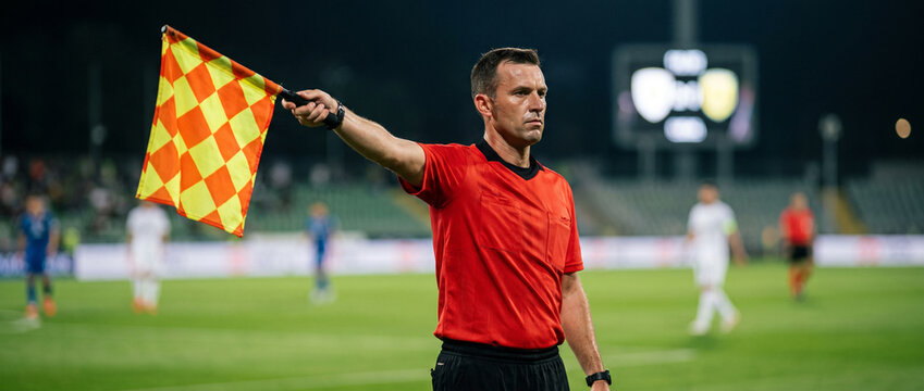 Linesman extends checkered flag to signal football match decision. Concept of offside control, referee teamwork and rules enforcement.