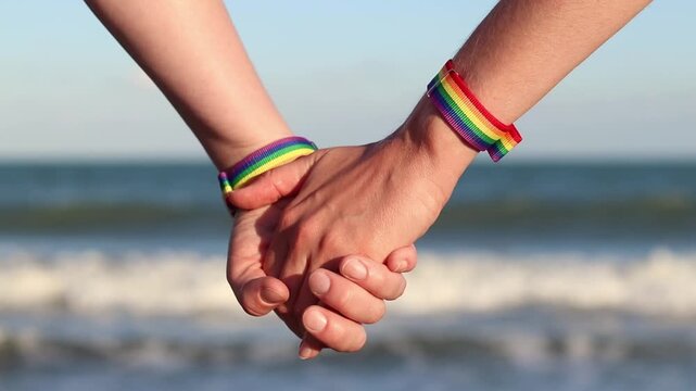 Same-sex couple with LGBTQ+ bracelets on their wrists. Two girls holding hands against the sea. Couple holding hands on the beach, showing off their pride bracelets. Love and equality concept