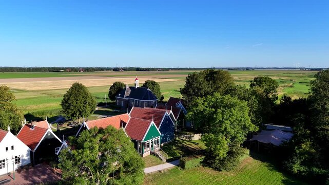 A scenic drone shot capturing the historic former island of Schokland in the Netherlands on a bright, sunny day. Pull back shot of church, revealing boat, with two cyclists.