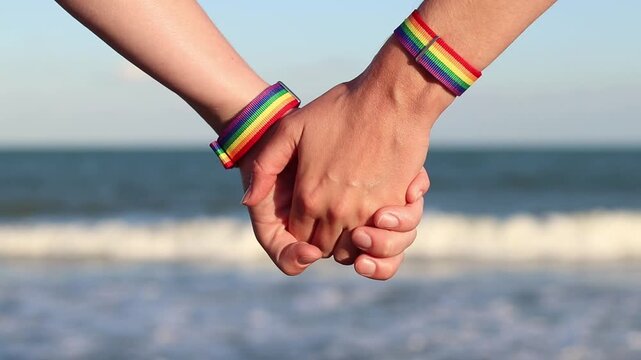 Same-sex couple with LGBTQ+ bracelets on their wrists. Two girls holding hands against the sea. Couple holding hands on the beach, showing off their pride bracelets. Love and equality concept