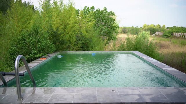An empty infinity pool sits surrounded by greenery and countryside fields, with a ladder and floating toys nearby, Static Shot
