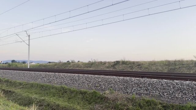 Blurred train passing by railway tracks with overhead power lines in a quiet countryside landscape
