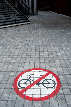 Urban pavement with a no bicycles sign marking a pedestrian passage where stairs and railing lead through a shadowed architecture exterior