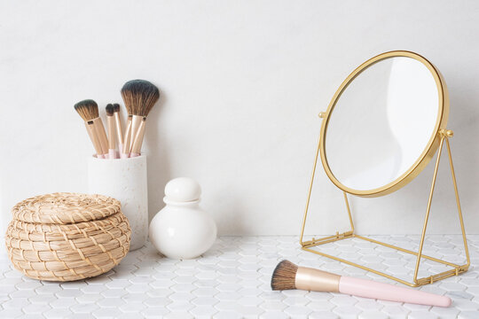 Holder with makeup brushes, cosmetics and mirror on white table close-up over light wall background