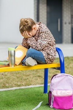 Sad Schoolgirl Sitting Alone with Book and Backpack Outdoors