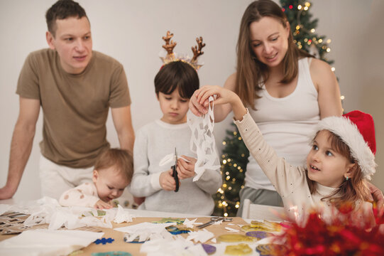 Happy family cut paper snowflakes near Christmas tree, celebrating Christmas with together. New Year's preparations and decorations. Paper crafts. 