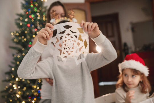 Happy family cut paper snowflakes near Christmas tree, celebrating Christmas with together. New Year's preparations and decorations. Paper crafts. Boy with paper snowflake